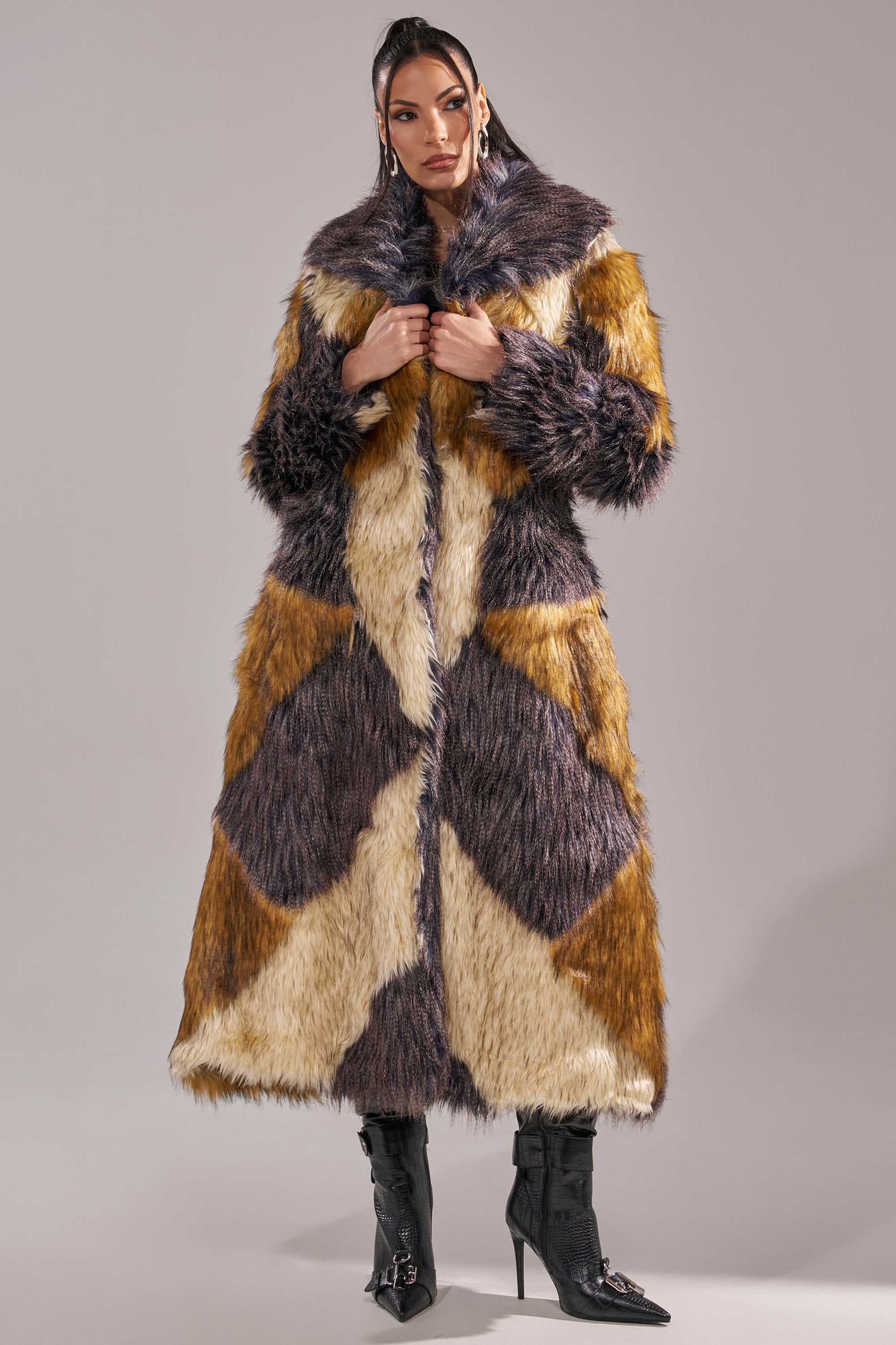A woman wears the NOTRE DAME FAUX FUR COAT in brown, tan, and gray patchwork over black pointed-toe heeled boots, standing against a plain gray background and capturing modern luxury street style.