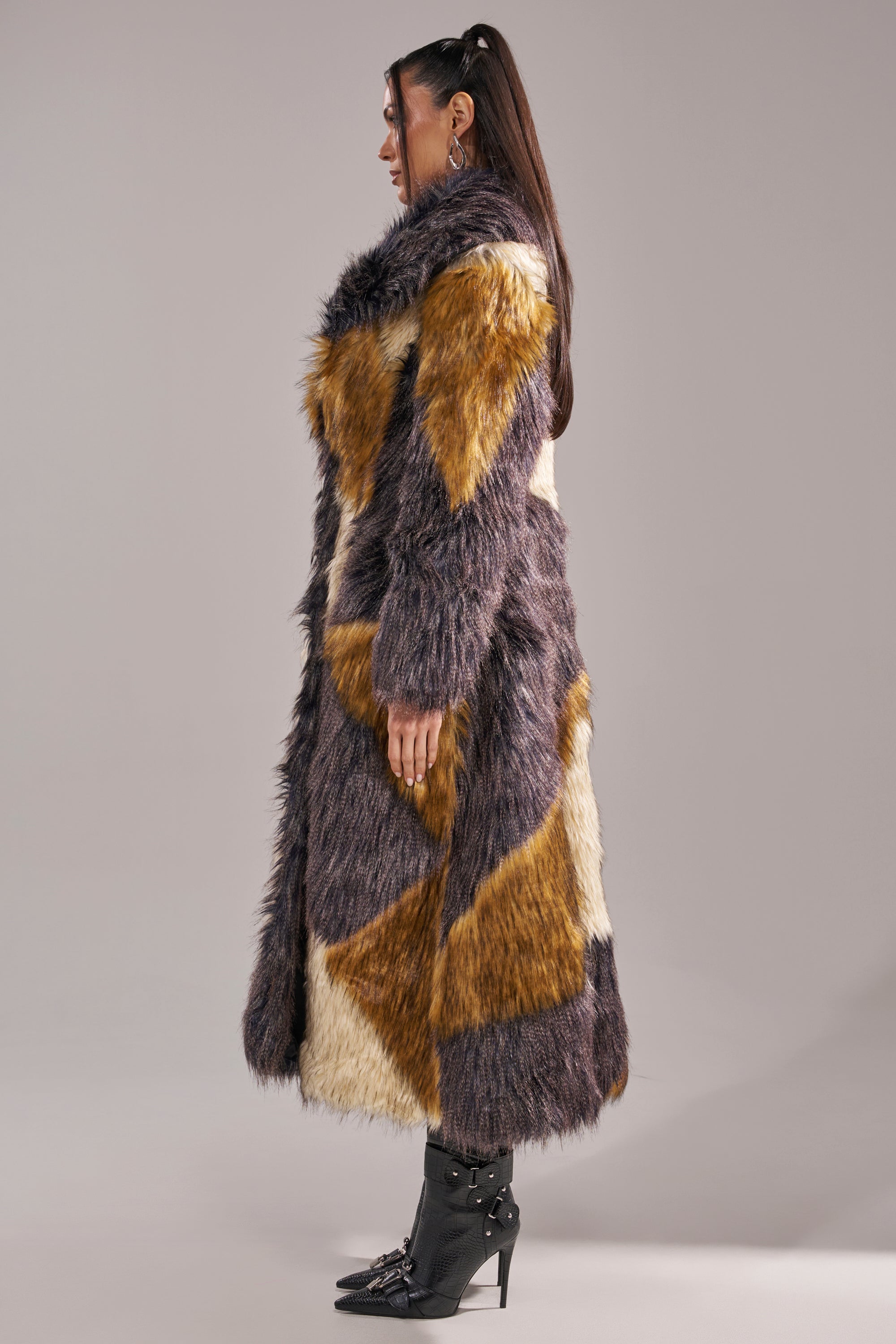 A woman stands in profile wearing the NOTRE DAME FAUX FUR COAT—a long, patchwork design in brown, cream, and gray—paired with black studded ankle boots for a bold luxury street style look against a neutral background.