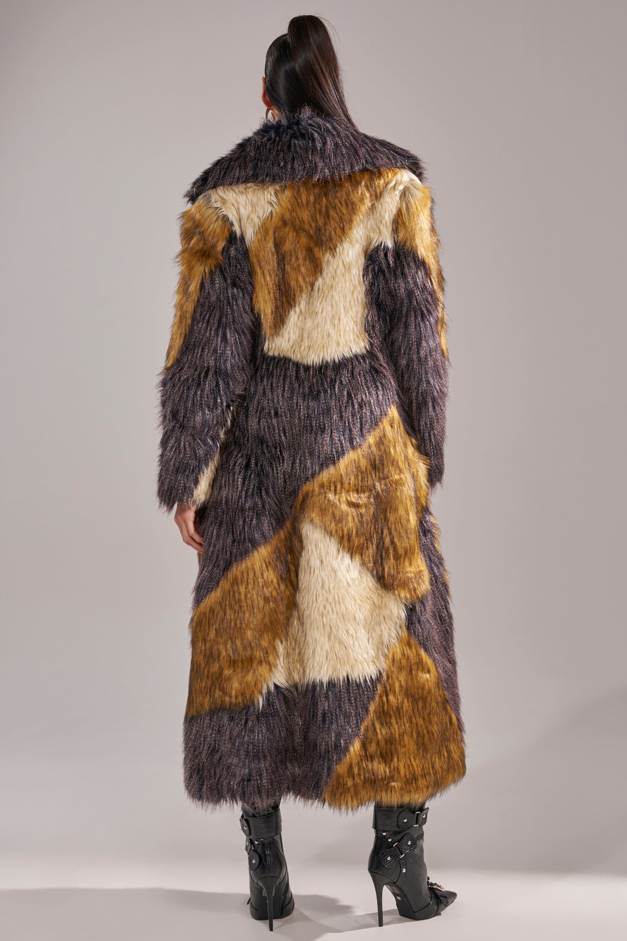 A person with long dark hair in a high ponytail models the NOTRE DAME FAUX FUR COAT, featuring a multicolored patchwork design in brown, cream, and dark tones, standing with their back to the camera against a plain background.