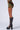 A person wearing a green and brown patterned IN THE DARK MINI SKIRT with black knee-high platform lace-up boots stands against a plain white background. The image captures the side profile, focusing on the lower half of their body to highlight the skirt's stretchable fabric.