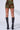 Rear view of a person wearing the IN THE DARK MINI SKIRT, featuring a green and brown floral ruched design with velvet floral accents, paired with black knee-high boots. The stretchy fabrication ensures a snug fit as the person stands with legs slightly apart against a plain white background.