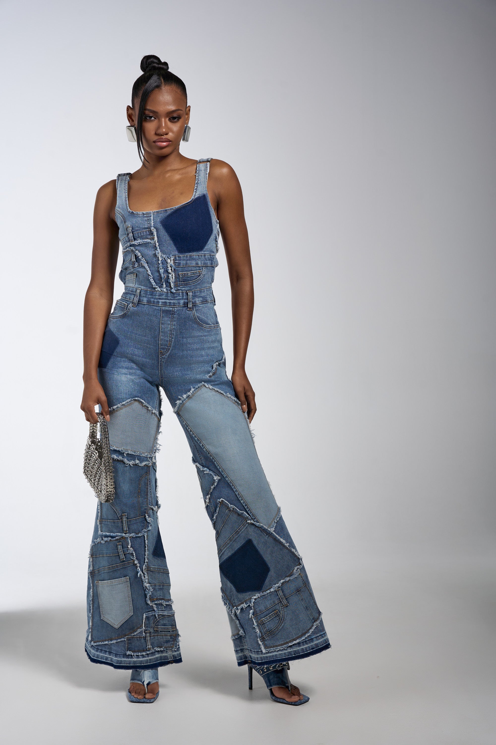 A woman poses confidently against a light background in the LIFE OF THE PARTY DENIM PATCHWORK JUMPSUIT with flared legs, paired with heeled sandals and a small silver handbag, her hair styled in a high ponytail.