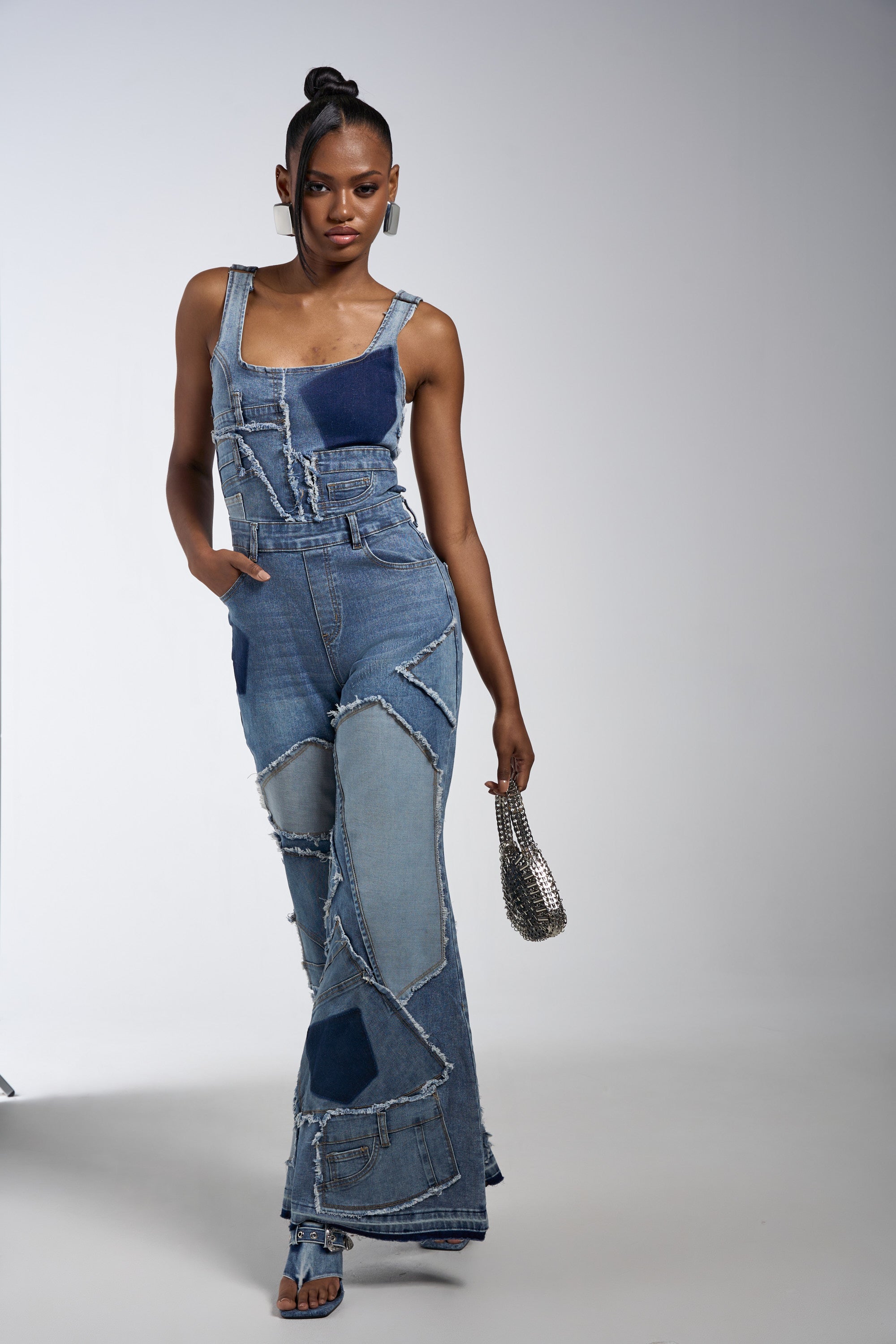 A woman models the LIFE OF THE PARTY DENIM PATCHWORK JUMPSUIT featuring deconstructed denim with frayed edges, one hand in her pocket, styled with high-heeled sandals, statement earrings, a silver handbag, and a topknot.