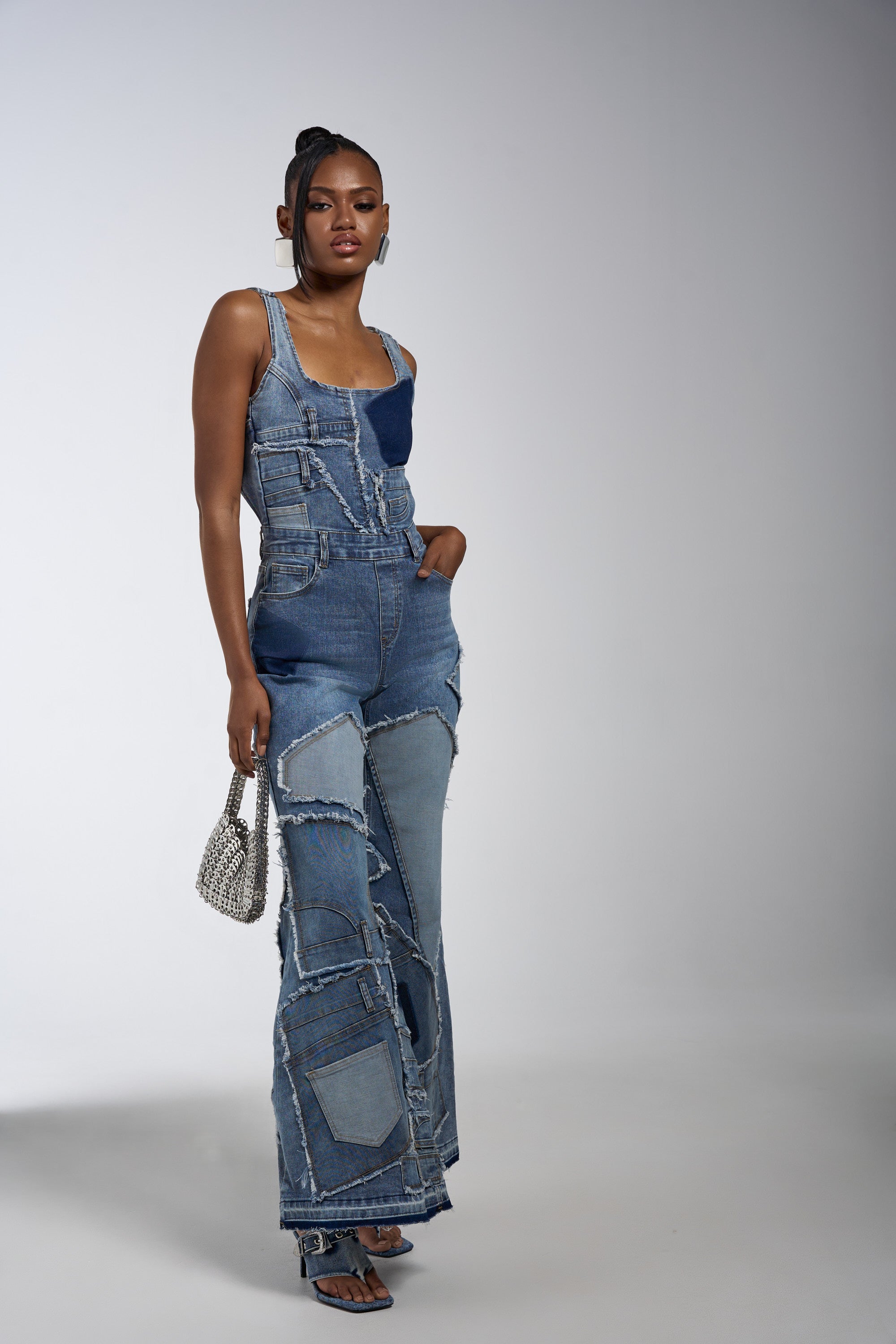 A woman models the LIFE OF THE PARTY DENIM PATCHWORK JUMPSUIT with frayed edges, paired with blue sandals and a sparkly handbag, standing against a plain gray background with one hand in her pocket.