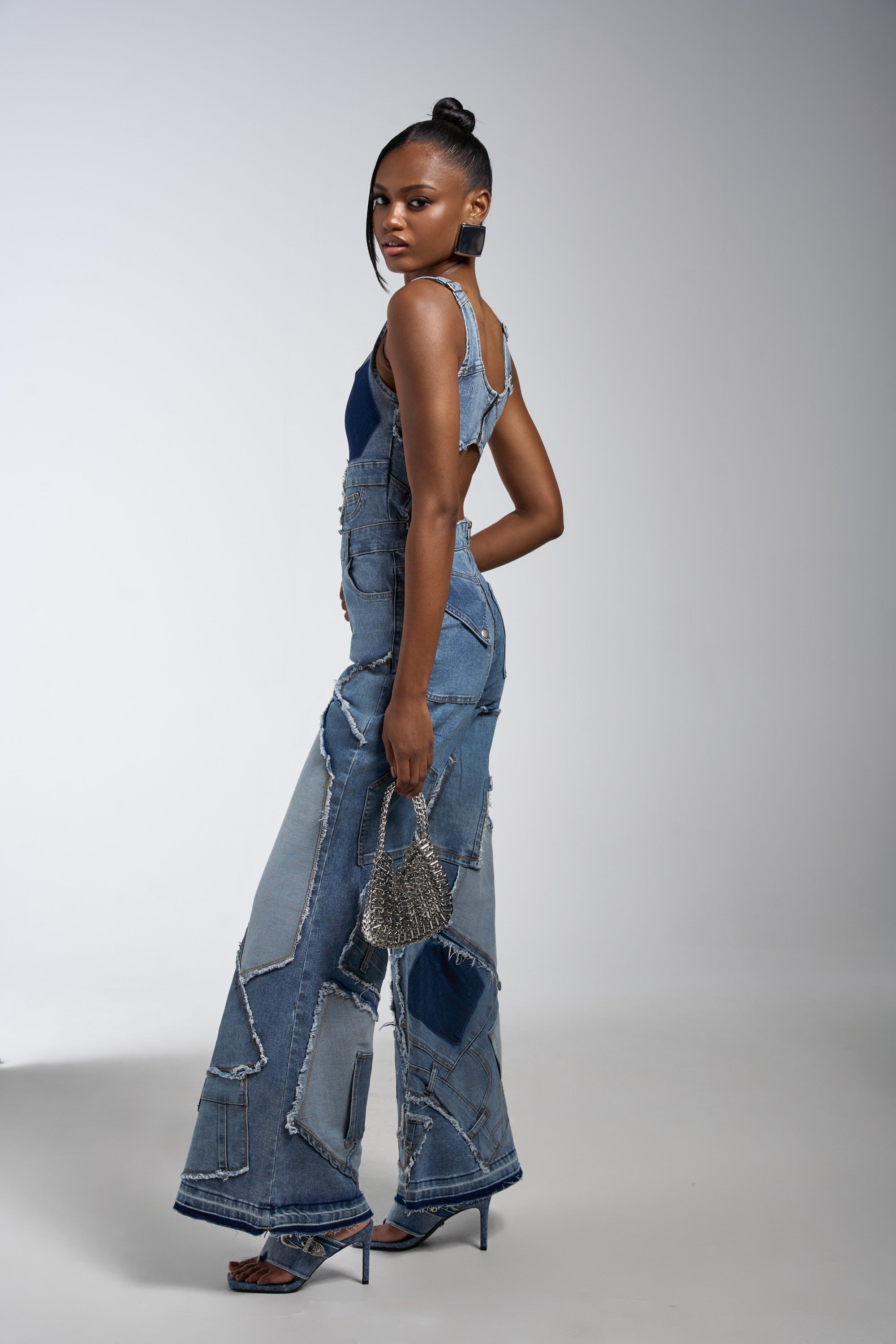 A woman poses in a studio wearing the LIFE OF THE PARTY DENIM PATCHWORK JUMPSUIT, styled with heeled sandals and a sparkling purse. She looks over her shoulder with a neutral expression against a plain light background.