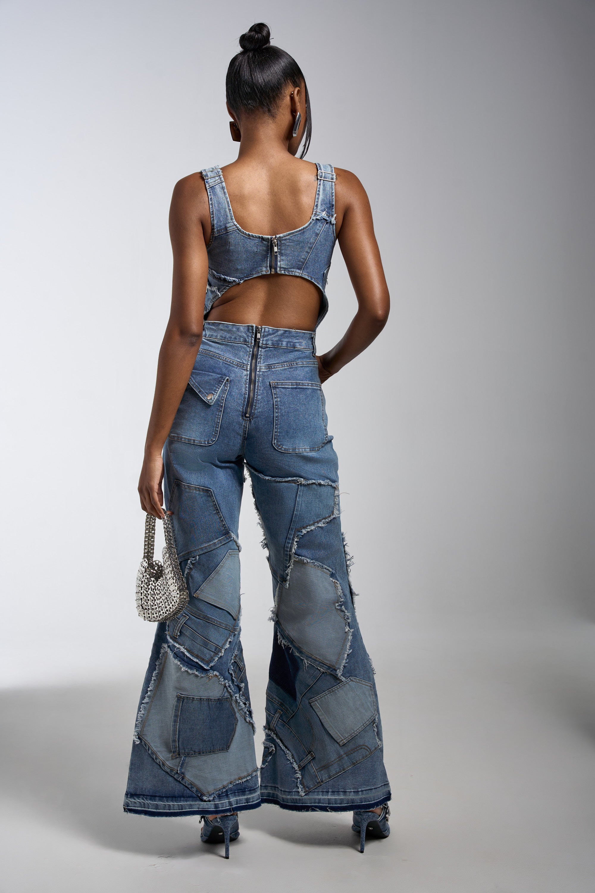 A woman faces away, modeling the LIFE OF THE PARTY DENIM PATCHWORK JUMPSUIT with flared legs and a sleeveless, back-cut design. She carries a silver handbag and wears high heels against a plain gray backdrop.