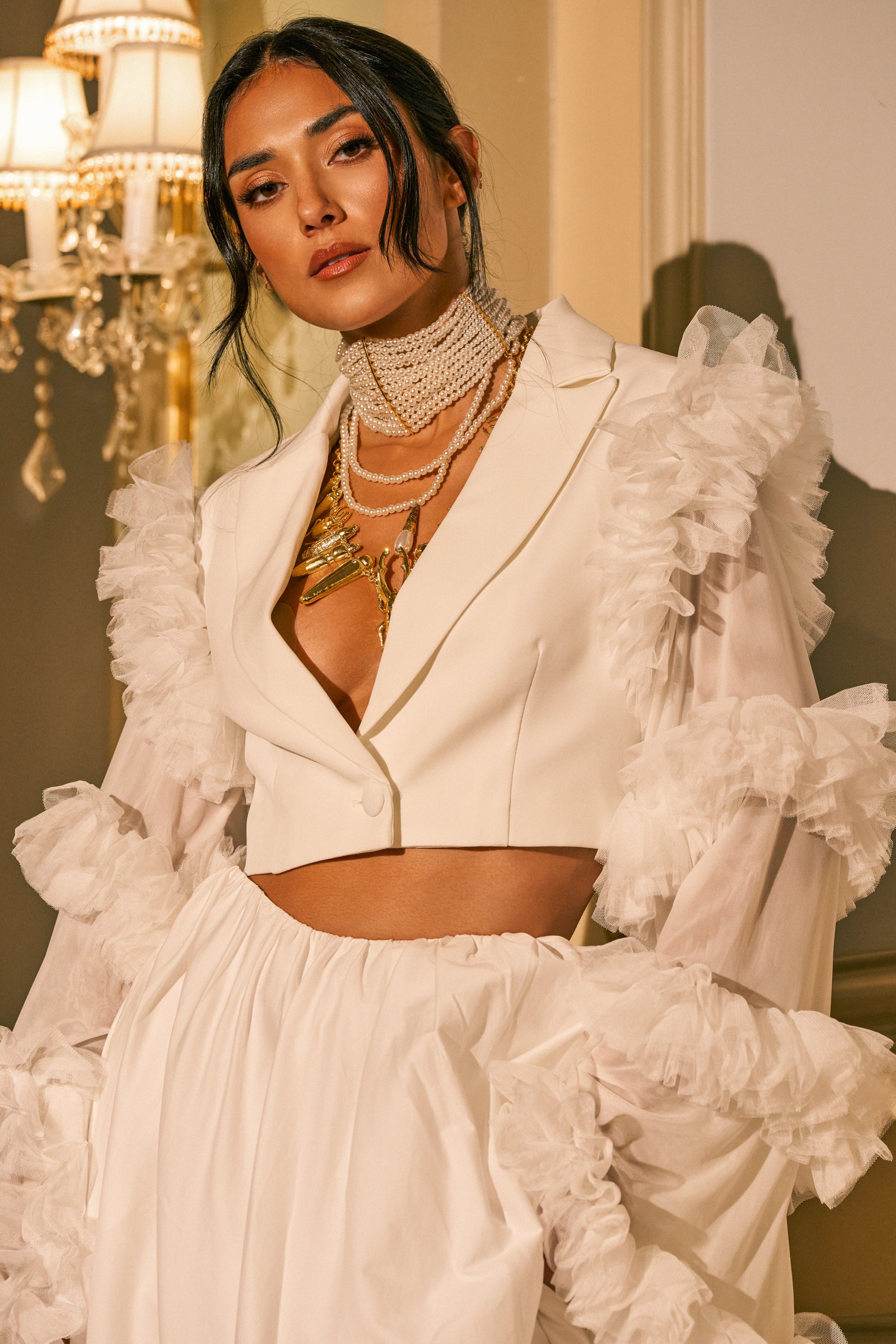 A woman poses indoors in the FLORA RUFFLE SLEEVE BLAZER, featuring stylish ruffled tulle sleeves, paired with a matching high-waisted skirt and accessorized with layered gold necklaces and a pearl choker. Elegant lighting and a chandelier illuminate the background.