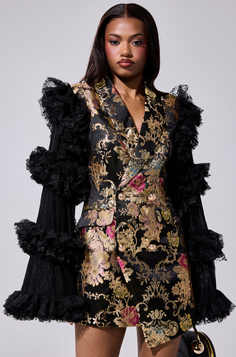 A woman in the LORY PRINTED BLAZER DRESS with black and gold floral brocade, ruffled lace sleeves, and long straight hair stands facing forward, carrying a small black handbag at her side.