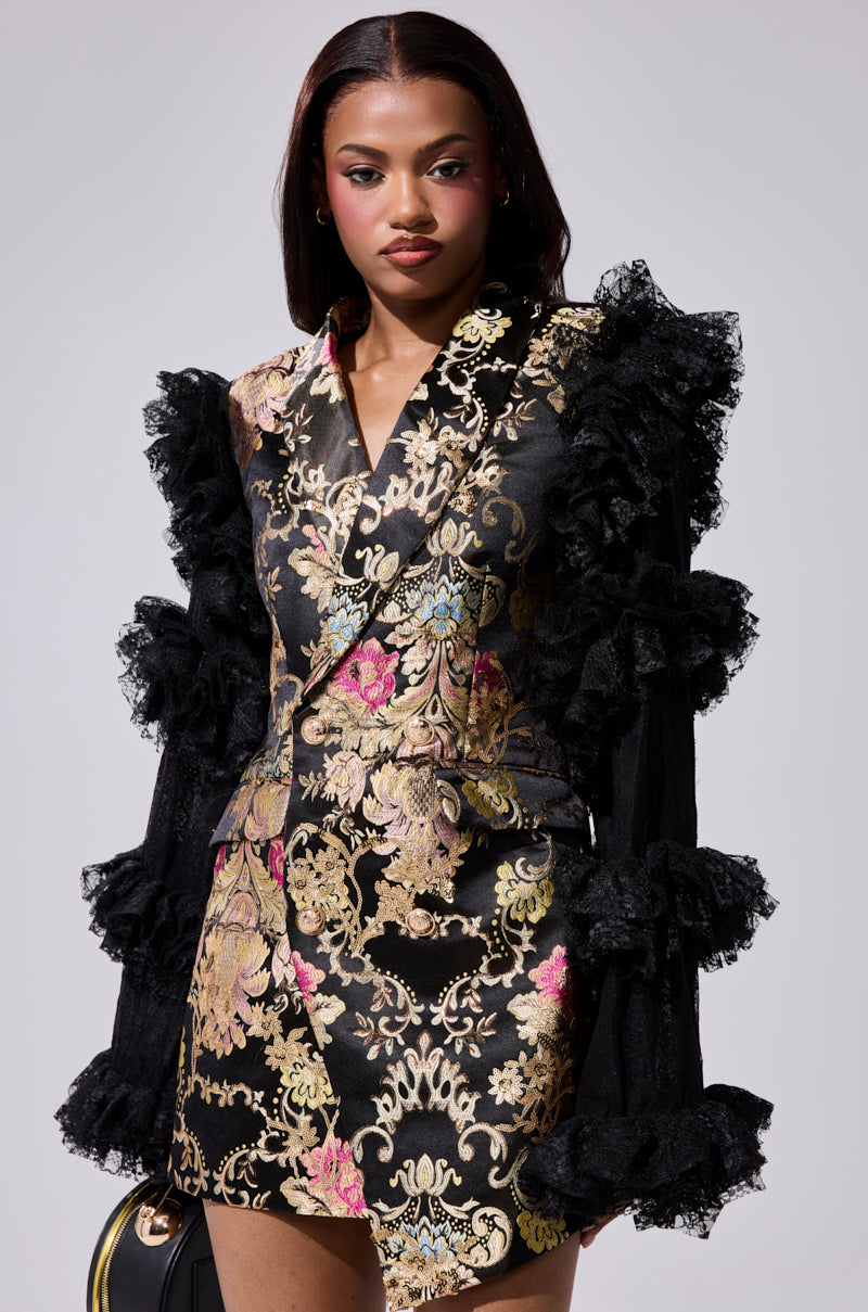 A woman models the LORY PRINTED BLAZER DRESS, featuring black and gold brocade with pink and gold floral patterns and dramatic black ruffled lace sleeves, against a plain light gray background.