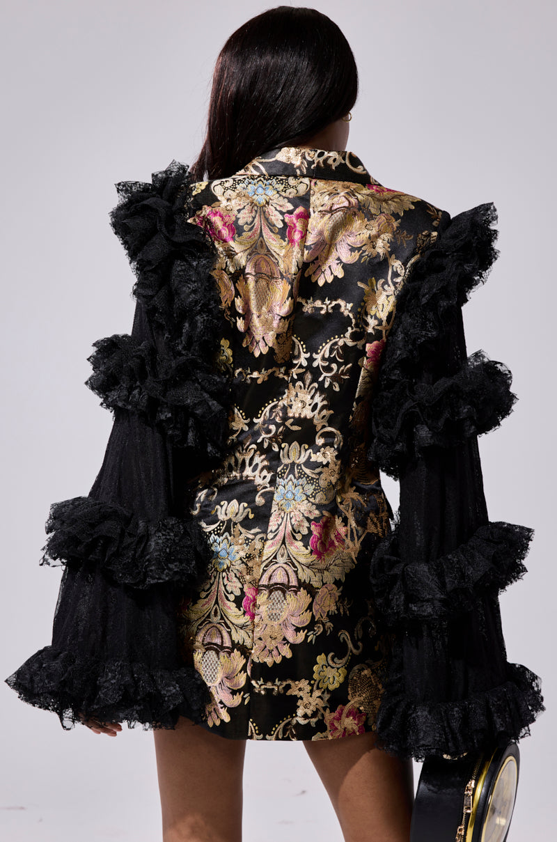 A person with long dark hair faces away, wearing the LORY PRINTED BLAZER DRESS featuring black and gold floral patterns, ornate scrollwork, and dramatic lace ruffles. They hold a round black bag against a plain background.