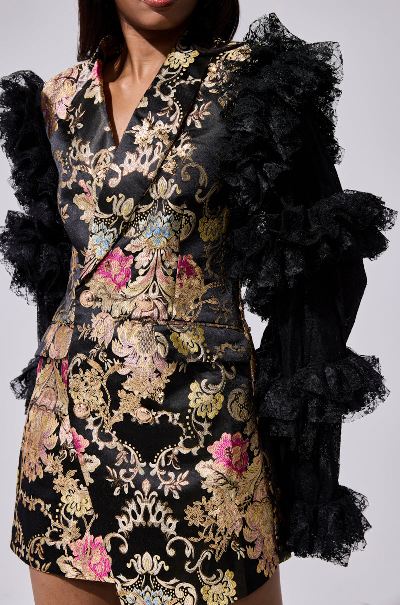 A woman models the LORY PRINTED BLAZER DRESS, featuring black brocade with gold and pink floral patterns and dramatic ruffled lace sleeves, shown from shoulders to mid-thigh against a plain background.