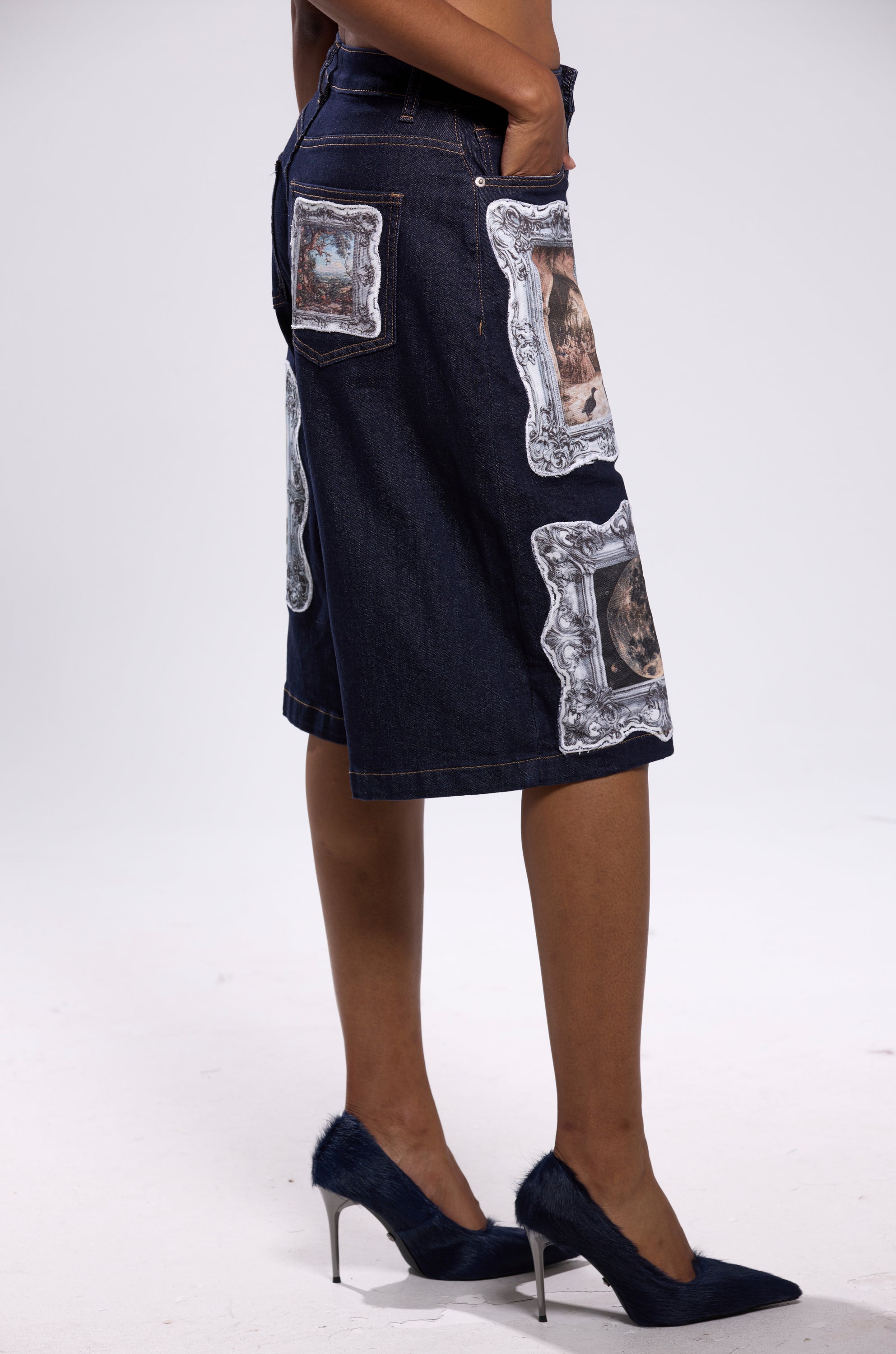 A person wears the PICTURE THIS BERMUDA SHORT—dark, vintage-inspired knee-length denim with picture frame graphics—and dark blue high heels, standing against a plain white background.