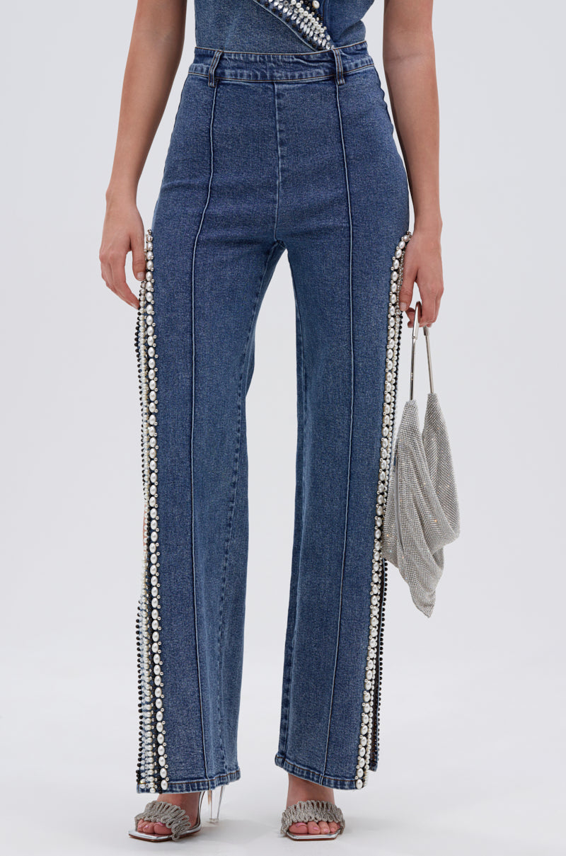 A person wears LUXURIOUS DENIM PANTS WITH SIDE SLITS embellished with pearls on the outer seams, a matching denim top, heeled sandals, and carries a glittery silver clutch against a plain white background.