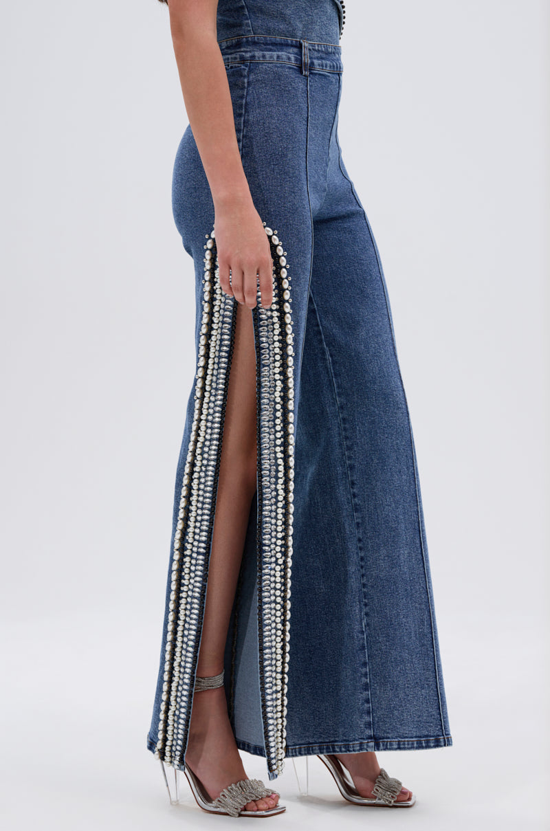 Only the lower body of a woman is visible, showcasing LUXURIOUS DENIM PANTS WITH SIDE SLITS adorned with pearls and rhinestones, paired with silver high-heeled sandals.