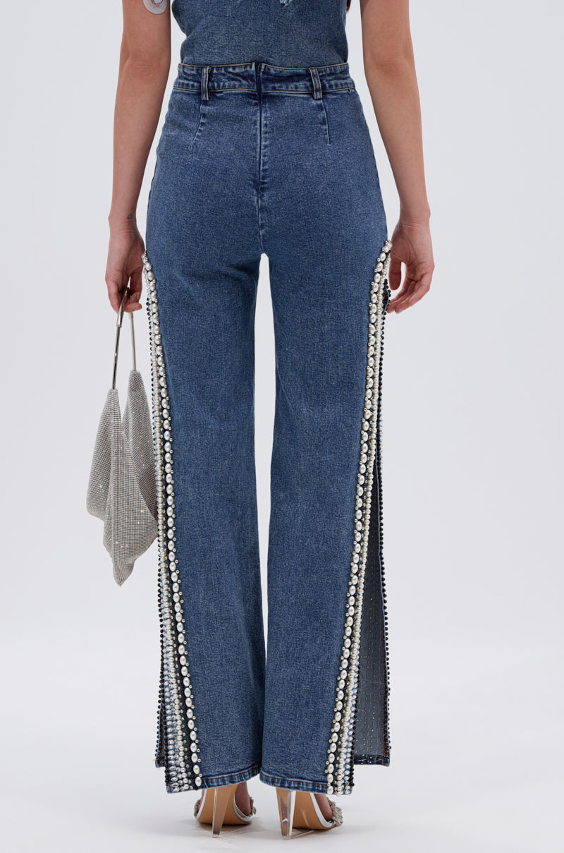 A person stands facing away, wearing LUXURIOUS DENIM PANTS WITH SIDE SLITS featuring pearl details and flared legs, paired with a sparkling silver handbag and clear high-heeled sandals for a chic finish.