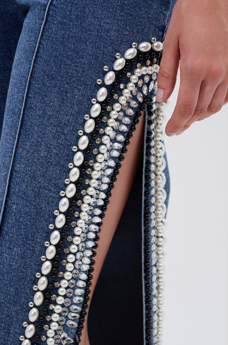 A close-up of a hand holding the open seam of LUXURIOUS DENIM PANTS WITH SIDE SLITS, showcasing pearl detailing and revealing skin underneath.