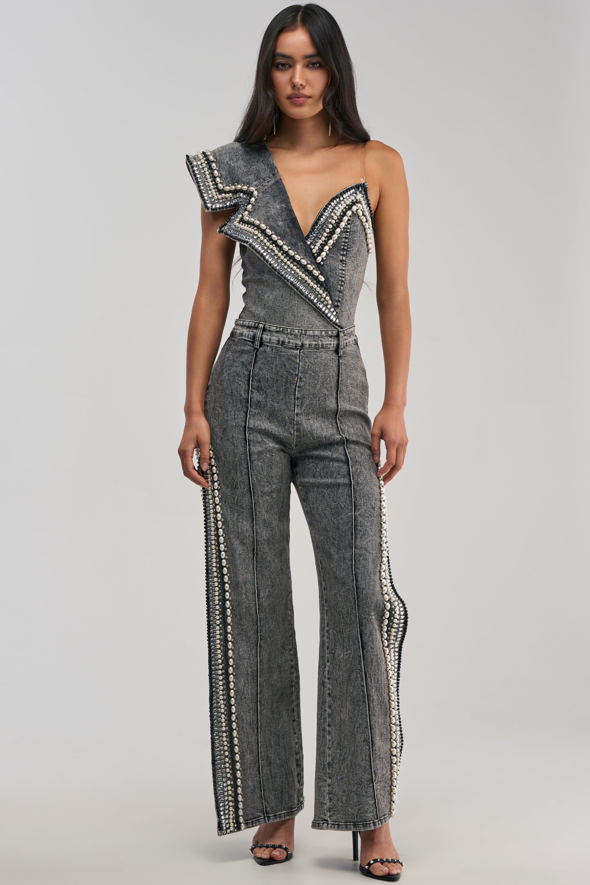 A woman stands against a plain background wearing LUXURIOUS DENIM PANTS WITH SIDE SLITS, styled with black high-heeled sandals and a chic grey, one-shoulder jumpsuit featuring pearl rhinestone embellishments. The pants have flared legs reminiscent of washed grey denim.