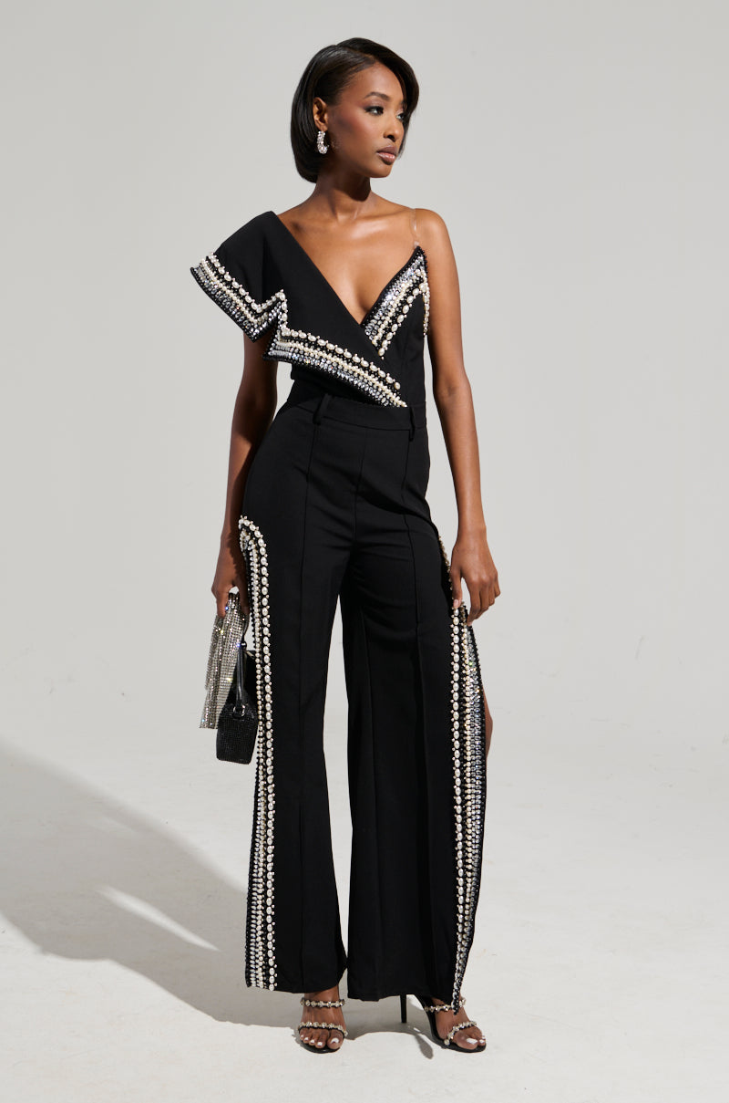 A woman in a stylish black jumpsuit adorned with rhinestones poses against a plain backdrop, showcasing luxurious pants with side slits and featuring a modern one-shoulder design. She complements the look with a silver clutch and strappy heels.