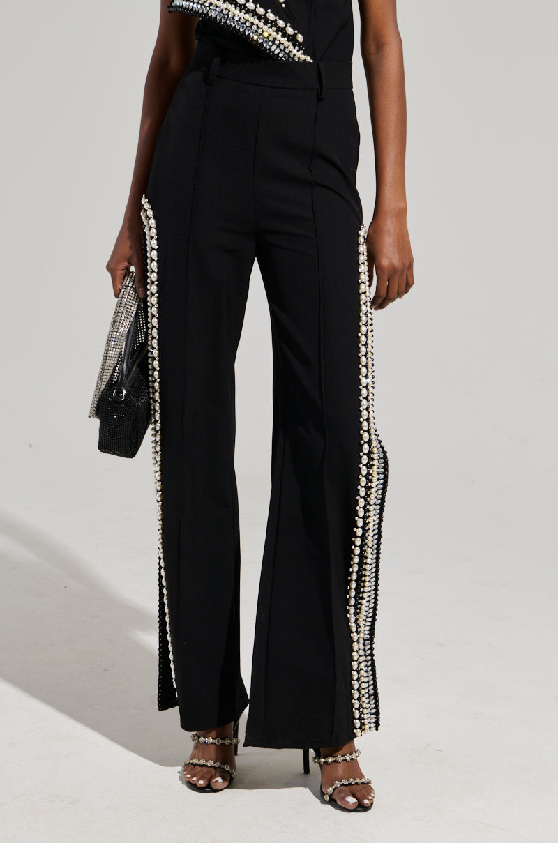 A person stands dressed in LUXURIOUS PANTS WITH SIDE SLITS in a rich black color, adorned with pearl-like beads and subtle side slits. They hold a shimmering black clutch and wear strappy high-heeled shoes, all set against a plain, light-colored background.