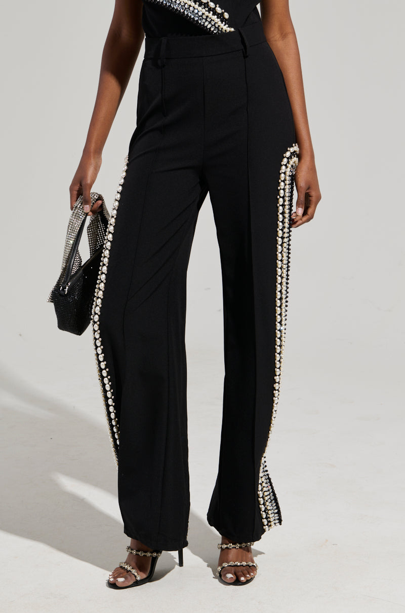 A person dressed in chic black LUXURIOUS PANTS WITH SIDE SLITS enhanced with rhinestones along the sides, beautifully paired with a black mesh top and silver-heeled sandals, carrying a small textured black handbag.