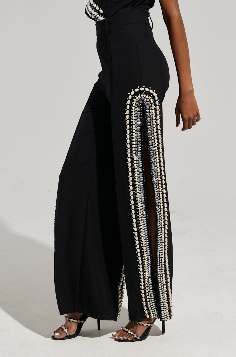 A person wearing the LUXURIOUS PANTS WITH SIDE SLITS in black, embellished with vertical rows of glistening beads, sequins, and rhinestones along the legs. The pants have a wide leg and feature side slits, paired with high-heeled sandals against a plain light-colored background.
