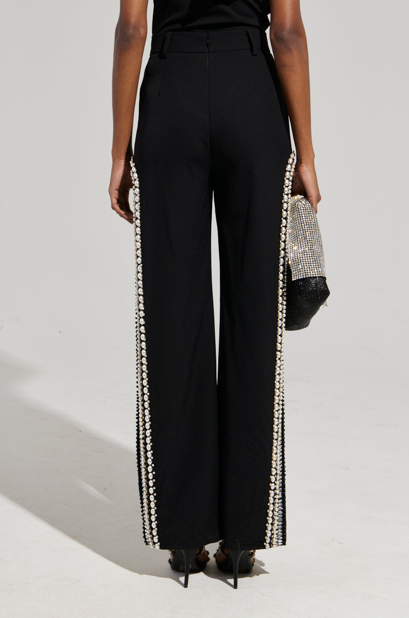 A person wearing high-waisted black LUXURIOUS PANTS WITH SIDE SLITS, adorned with opulent rhinestone embellishments along the sides, stands with their back to the camera, holding a small, sparkly handbag.