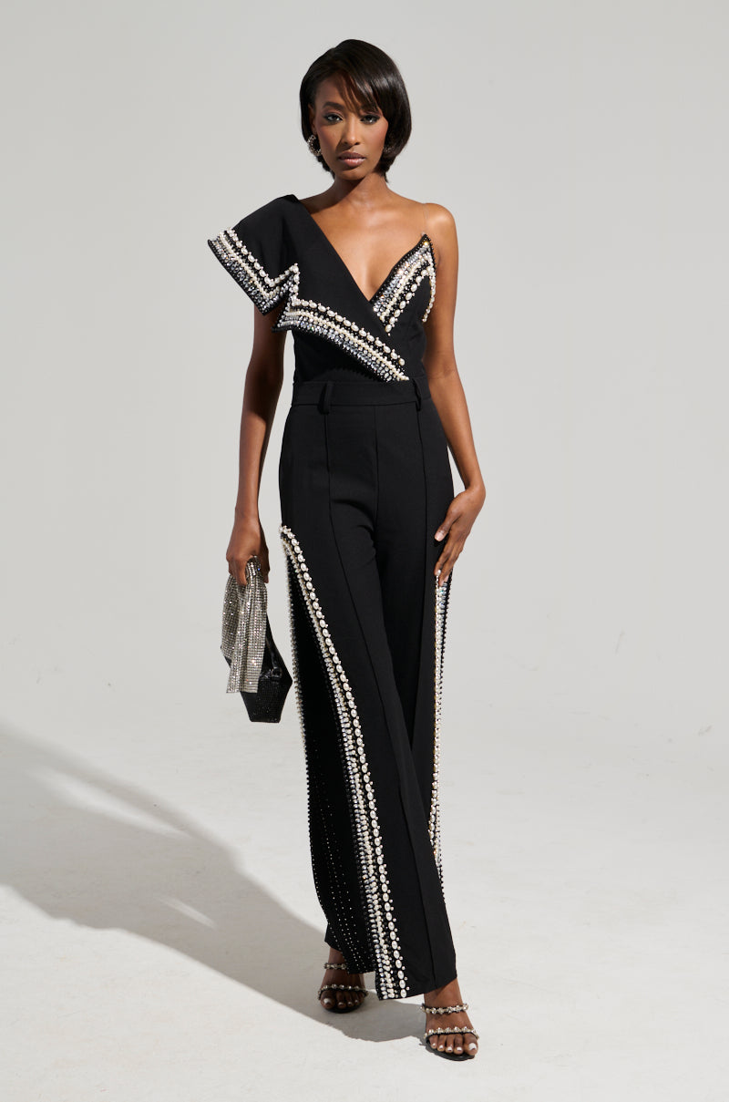 A woman in an elegant asymmetrical black ensemble detailed with rhinestones poses against a plain backdrop. She accessorizes with a glittering silver clutch and pairs her outfit with coordinating black-and-white beaded sandals. Her sleek bob hairstyle flawlessly complements the luxurious pants featuring side slits.