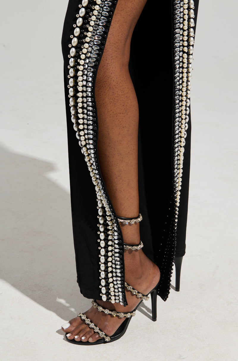 Close-up of a person wearing luxurious black pants with side slits, decorated with pearl and crystal embellishments. The matching stiletto heels and ankle strap complete the look, accentuating the elegant design.