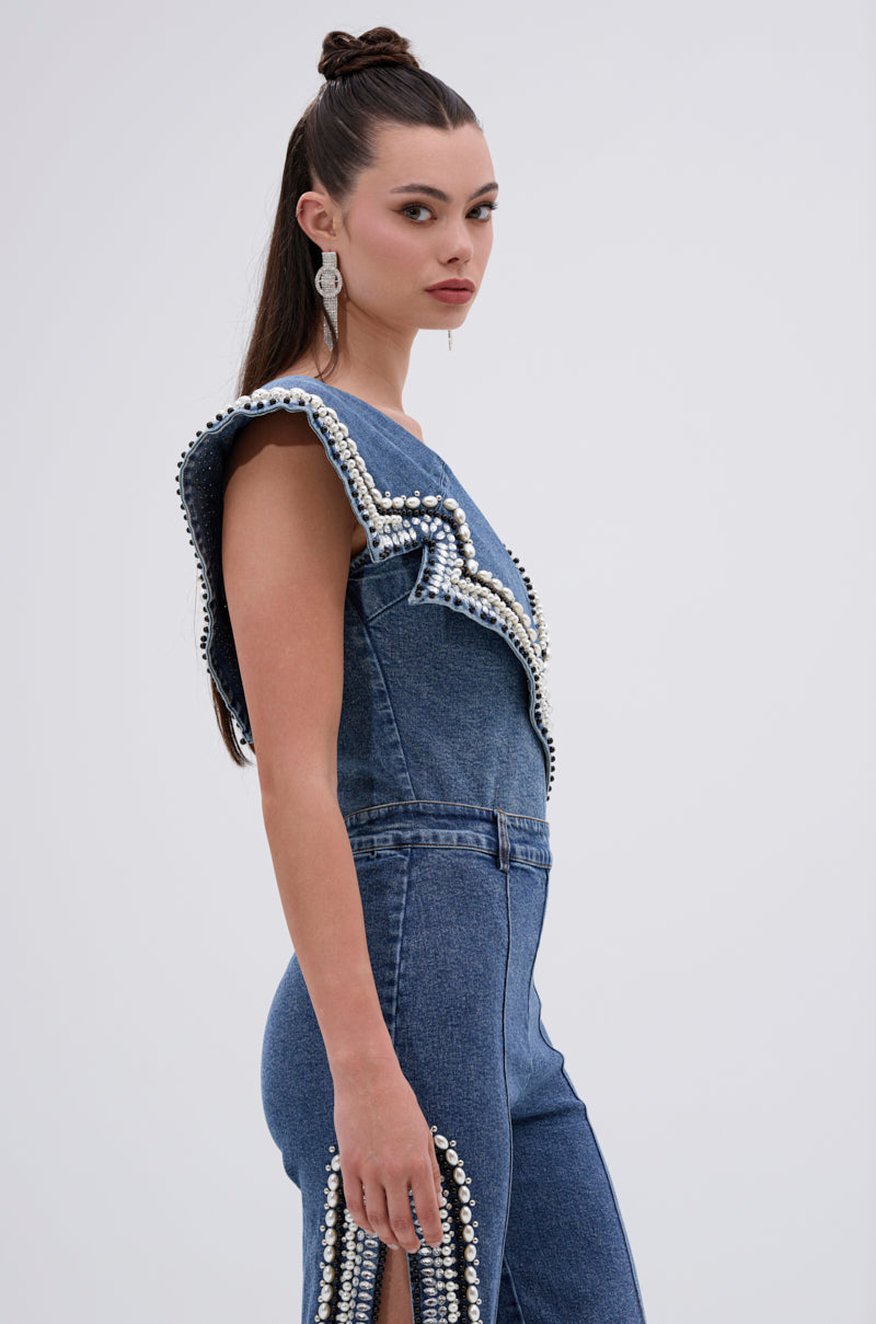 A woman with long, dark hair styled half up wears the LUXURIOUS DENIM ASYMMETRICAL PEARL BODYSUIT, featuring sleeveless denim and pearl-like bead accents. She faces sideways at the camera, accessorized with large silver earrings.