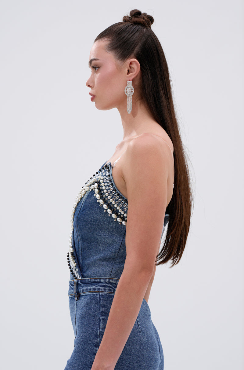 Woman in profile wearing the LUXURIOUS DENIM ASYMMETRICAL PEARL BODYSUIT with matching high-waisted jeans, long straight hair, and statement earrings, posed against a plain white background.