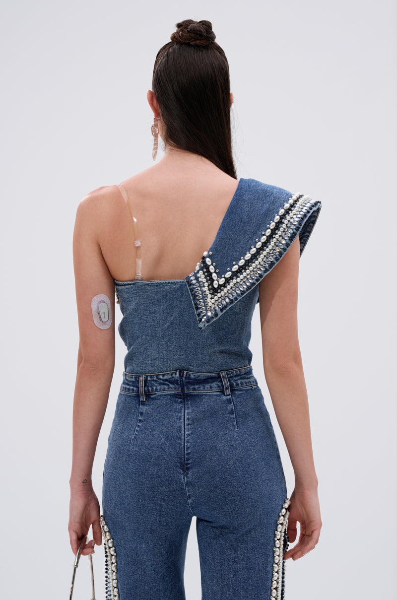 A woman faces away wearing the LUXURIOUS DENIM ASYMMETRICAL PEARL BODYSUIT with silver details and high-waisted jeans, a medical device on her upper arm, and a visible transparent bra strap.