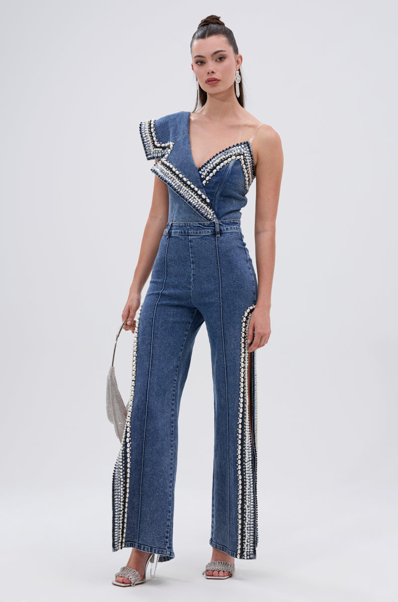 A woman poses against a plain background wearing the LUXURIOUS DENIM ASYMMETRICAL PEARL BODYSUIT, featuring pearl and studded embellishments with a one-shoulder ruffle. She carries a silver handbag and wears heeled sandals.