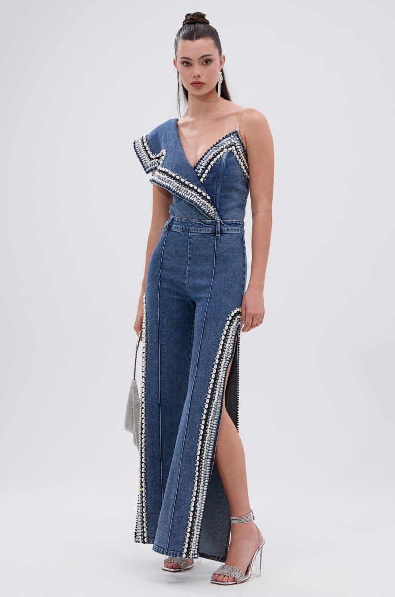 A woman with long dark hair in a high ponytail models the LUXURIOUS DENIM ASYMMETRICAL PEARL BODYSUIT, featuring wide legs, decorative trim, an asymmetrical one-shoulder neckline with pearl accents, and silver high-heeled sandals.