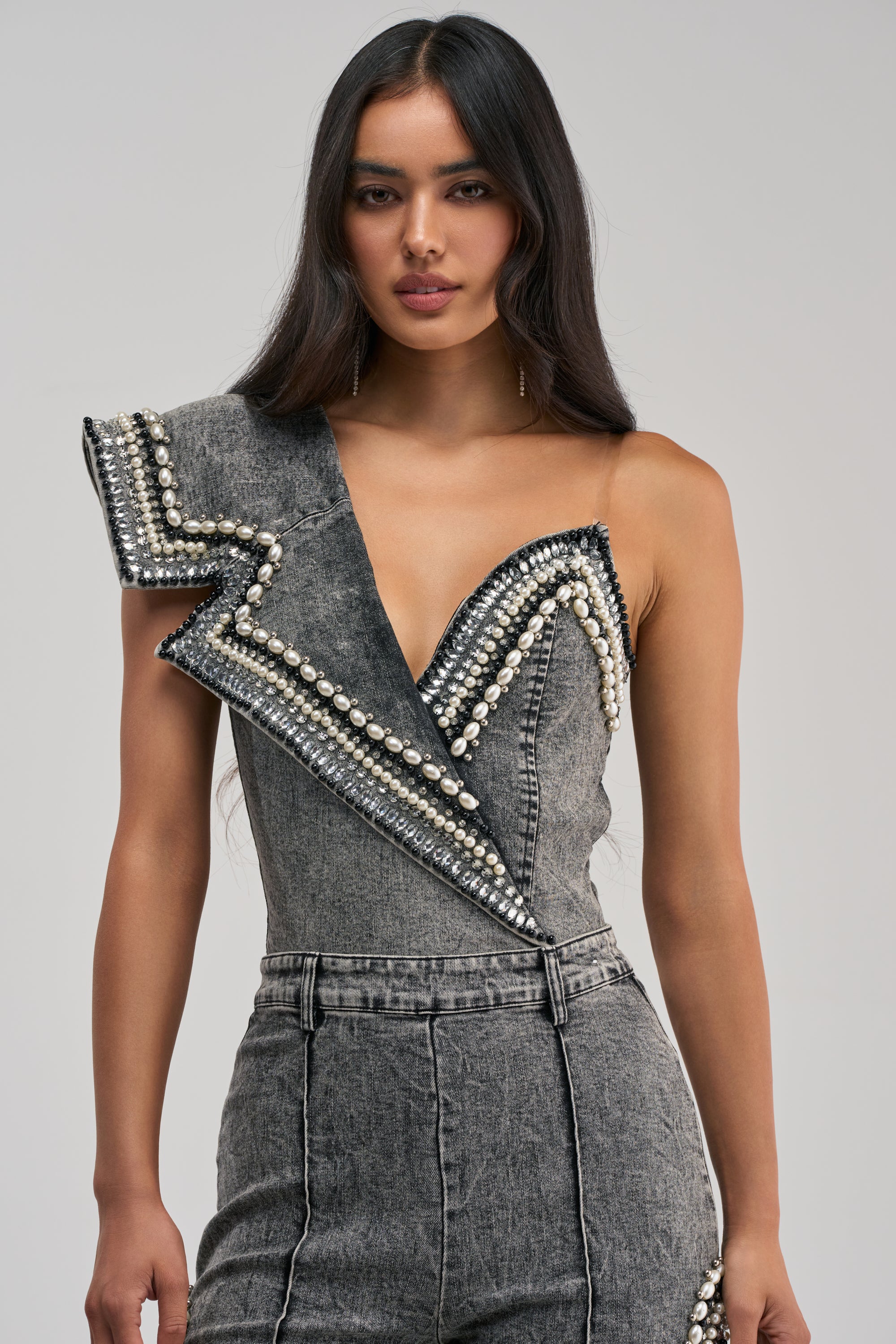 A woman with long dark hair models the LUXURIOUS DENIM ASYMMETRICAL PEARL BODYSUIT, featuring pearl and stud embellishments and an asymmetrical collar, against a plain background.