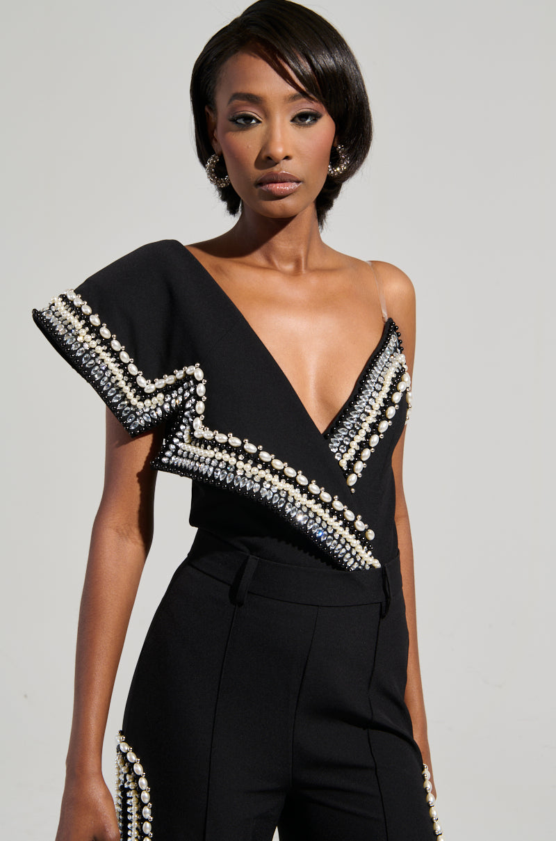 A woman dressed in the Luxurious Asymmetrical Pearl Bodysuit, featuring a chic black design with rhinestone accents and an elegant asymmetrical neckline and sleeve, is posed against a simple backdrop, radiating sophistication.