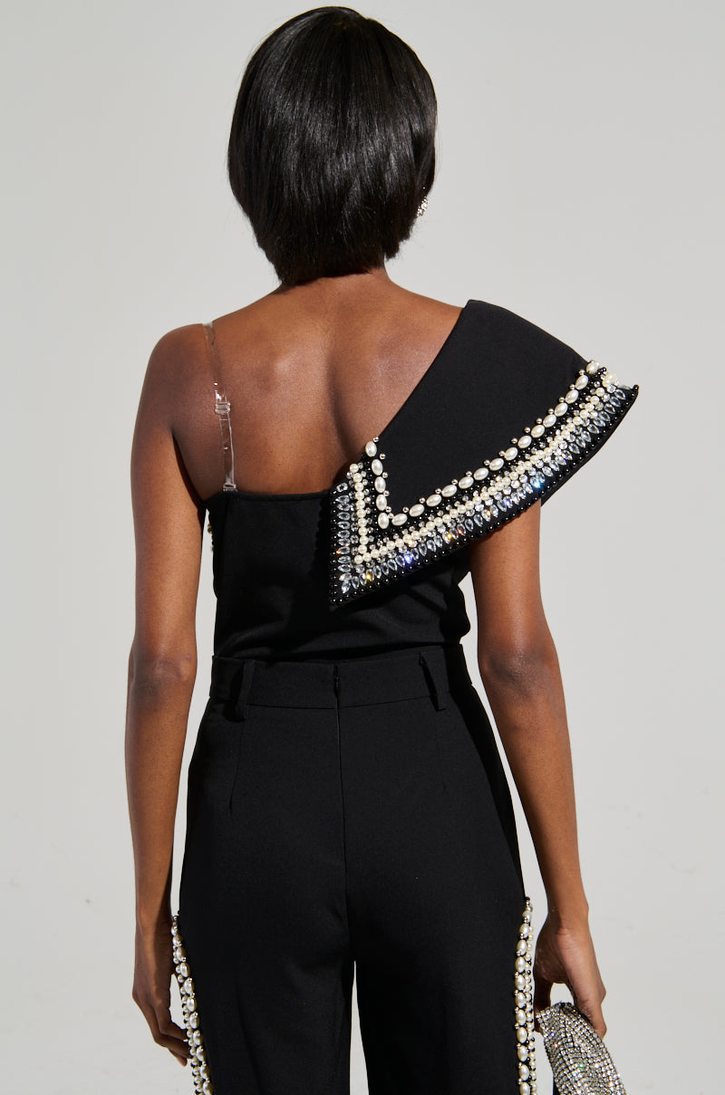 A person faces away from the camera, wearing the LUXURIOUS ASYMMETRICAL PEARL BODYSUIT in black, which highlights a rhinestone-embellished shoulder design. In one hand, they carry a textured clutch against a plain and neutral background.