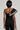 A person faces away from the camera, wearing the LUXURIOUS ASYMMETRICAL PEARL BODYSUIT in black, which highlights a rhinestone-embellished shoulder design. In one hand, they carry a textured clutch against a plain and neutral background.