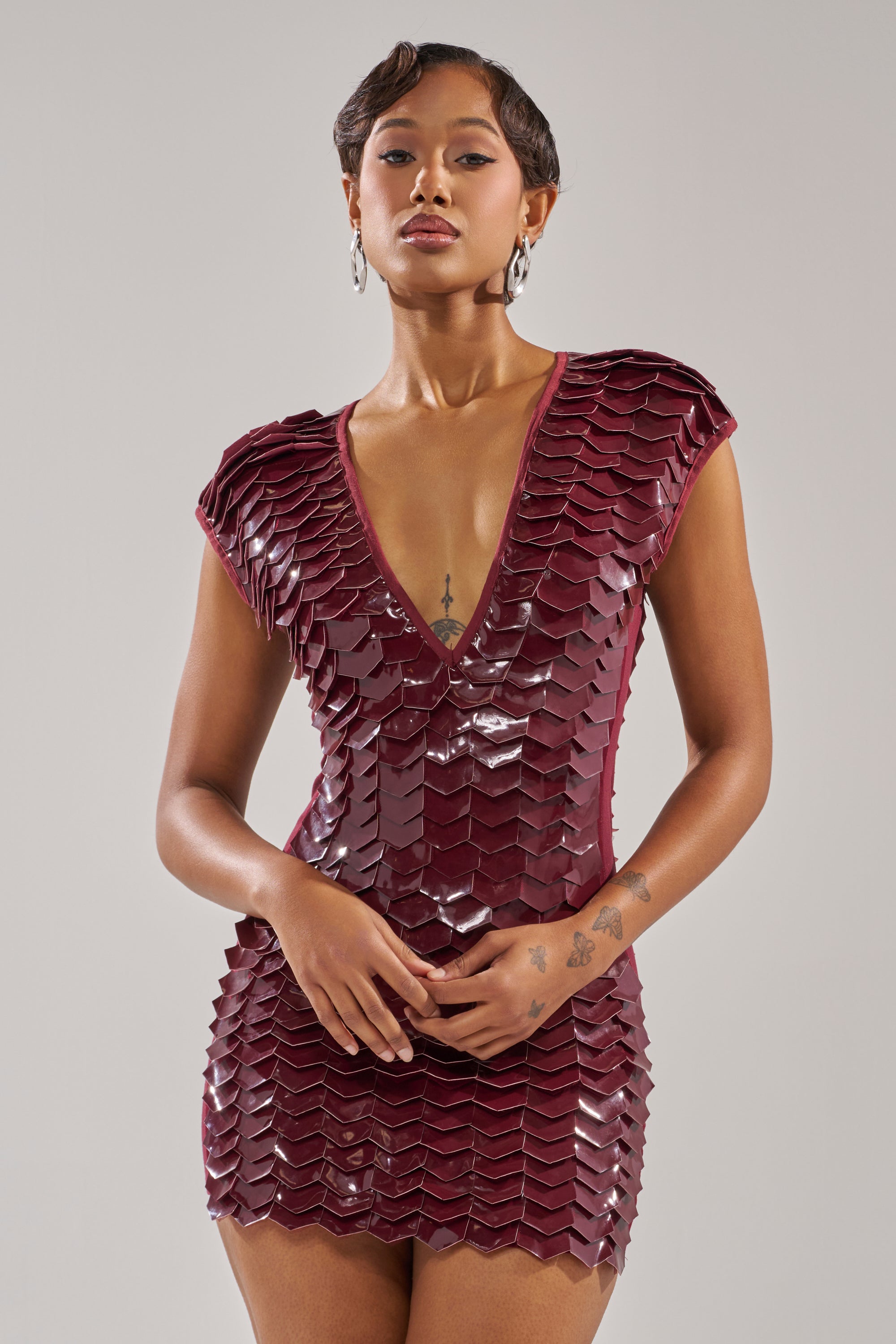 Wearing the CRAZY IN LOVE SCALE MINI DRESS in deep burgundy with a scale pattern and plunging neckline, a person stands against a plain background. Their tattoos, short hair, and hoop earrings add to the bold style.