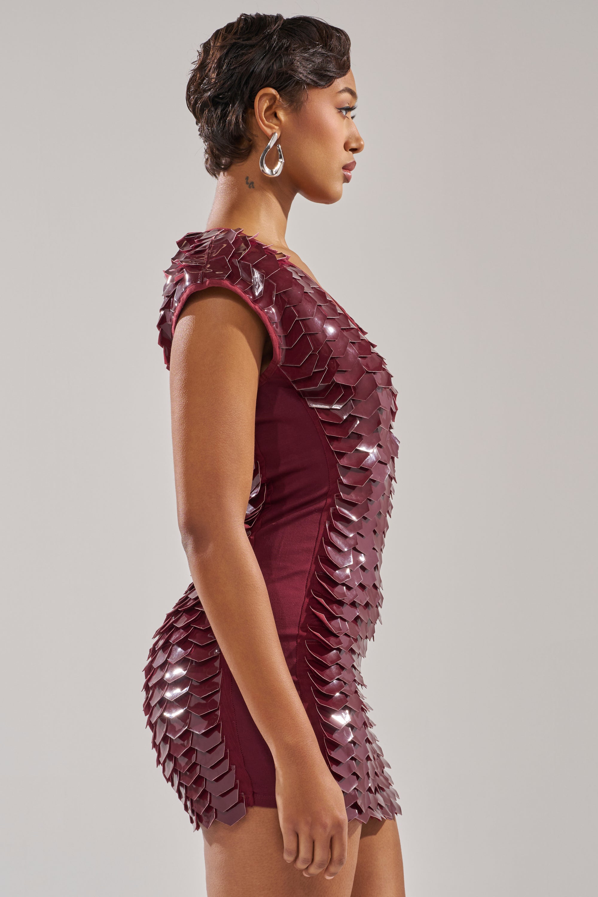 A woman in profile wears the CRAZY IN LOVE SCALE MINI DRESS, a fitted maroon bodycon mini with shiny, scale-like embellishments. She has short hair, hoop earrings, and stands against a plain light background.