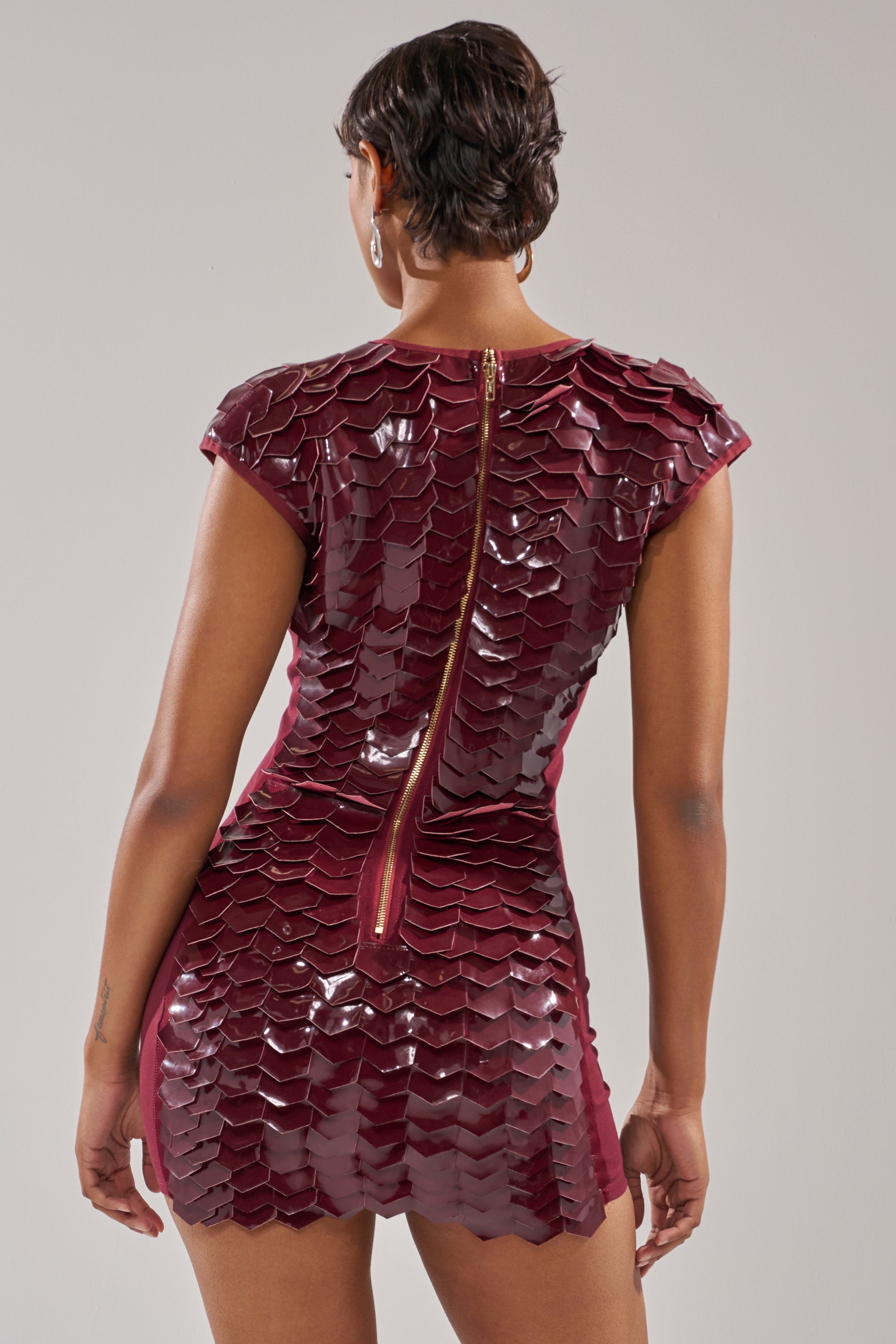 A person stands facing away, wearing the CRAZY IN LOVE SCALE MINI DRESS—a burgundy faux patent leather dress featuring shiny, scale-textured patterns and a visible gold zipper down the back.