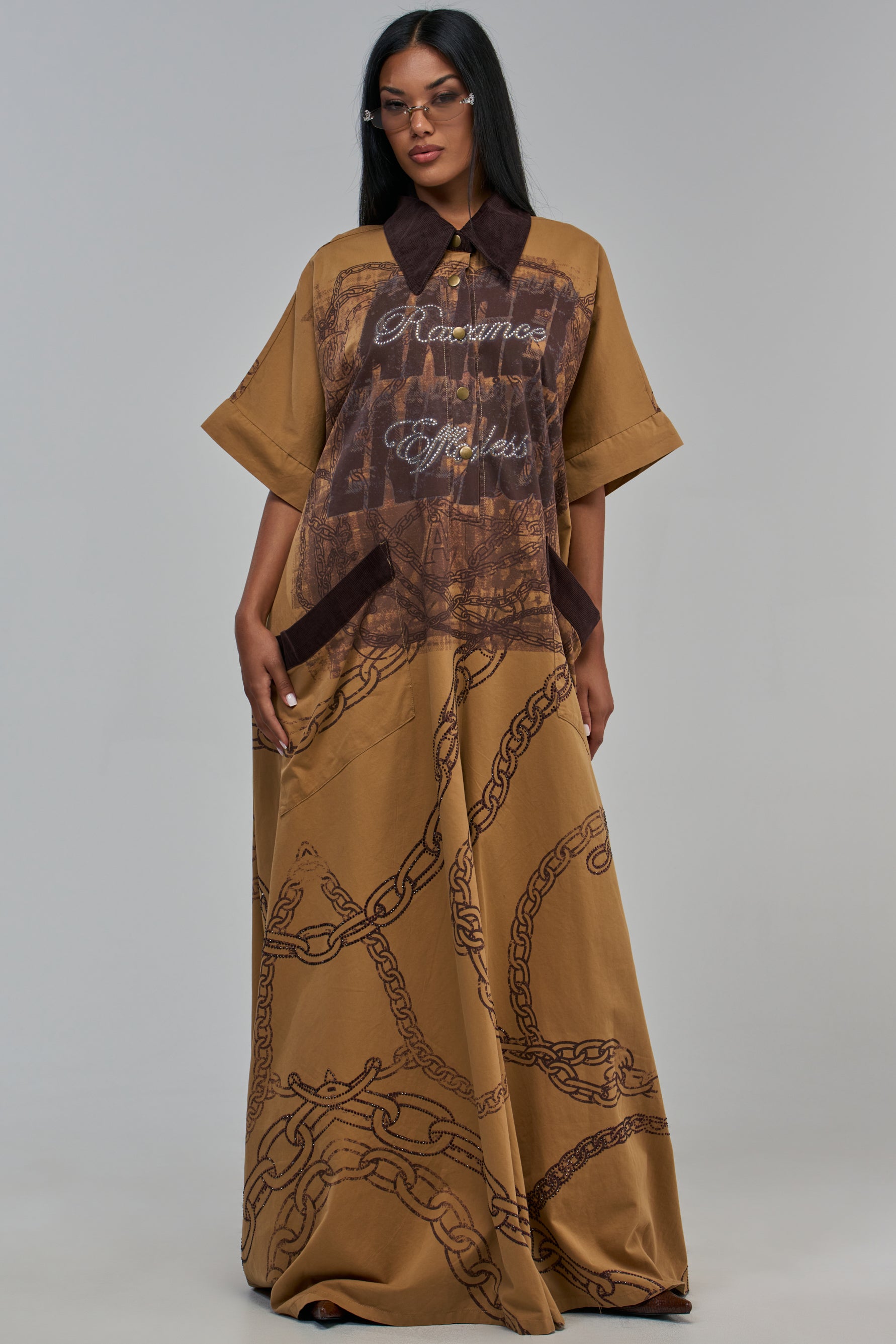 SHE’S RICH OVERSIZED EMBELLISHED JUMPSUIT IN TAN, an oversized khaki jumpsuit featuring a collared neckline, rhinestone embellishments on the chest, and a dark brown chain-link print on the skirt.