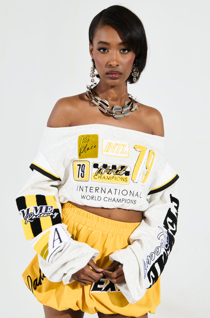 A woman radiates confidence in the RACING THROUGH OFF THE SHOULDER SWEATSHIRT, featuring racing-inspired graphics and text such as "1st Place" and "World Champions." She pairs it with a yellow skirt and a chunky necklace, standing elegantly against a plain backdrop. 1