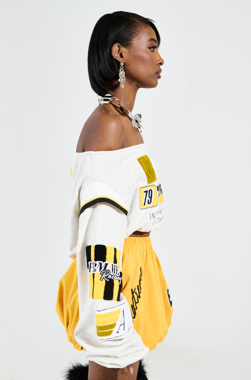 A woman with short, dark hair wears the chic RACING THROUGH OFF THE SHOULDER SWEATSHIRT adorned with racing-inspired graphics. She complements her look with high-waisted yellow shorts and hoop earrings, posing against a simple backdrop. 3