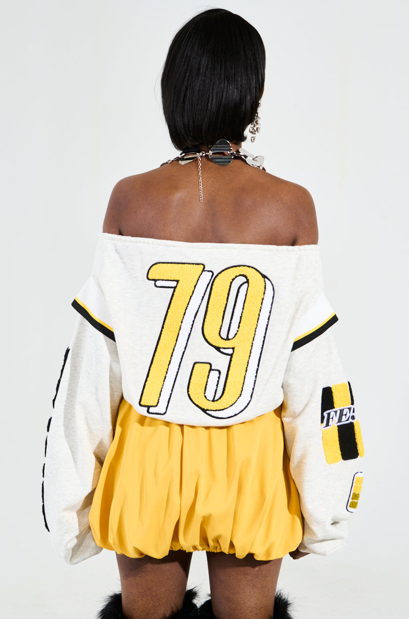 A person is captured from behind wearing the RACING THROUGH OFF THE SHOULDER SWEATSHIRT, which showcases racing-inspired graphics with the number 79 and a distinctive yellow and black pattern on the sleeve. They pair it with a yellow skirt and a necklace adorned with multiple chains and circular elements, while their hair is straight and falls to shoulder-length. 4