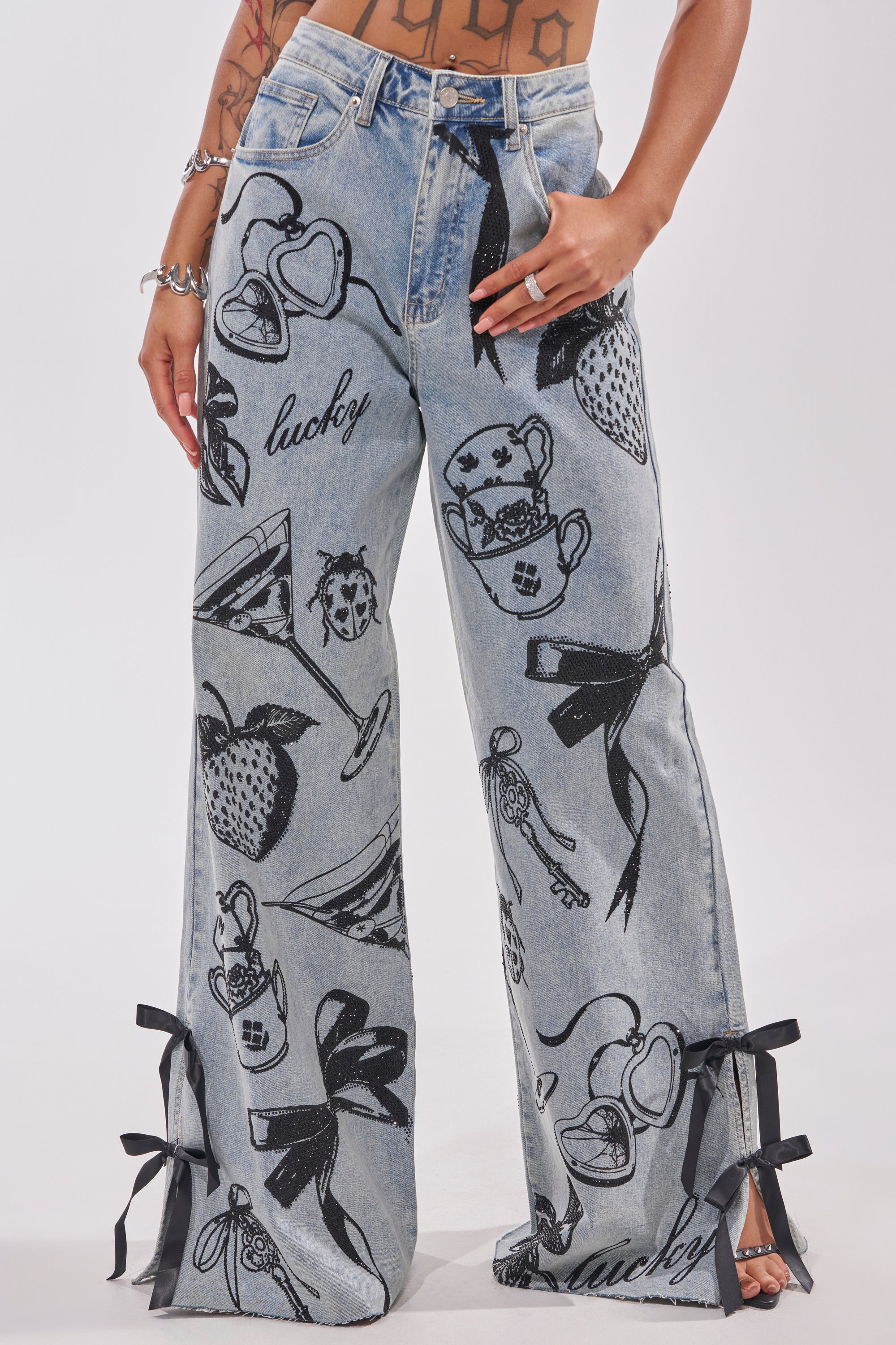 Someone wears AFTER HOURS STRAIGHT LEG DENIM, light blue jeans featuring black sketch graphics of hearts, strawberries, ladybugs, teacups, ribbons, and "lucky," with black ribbon bows accenting each leg's hem. 1