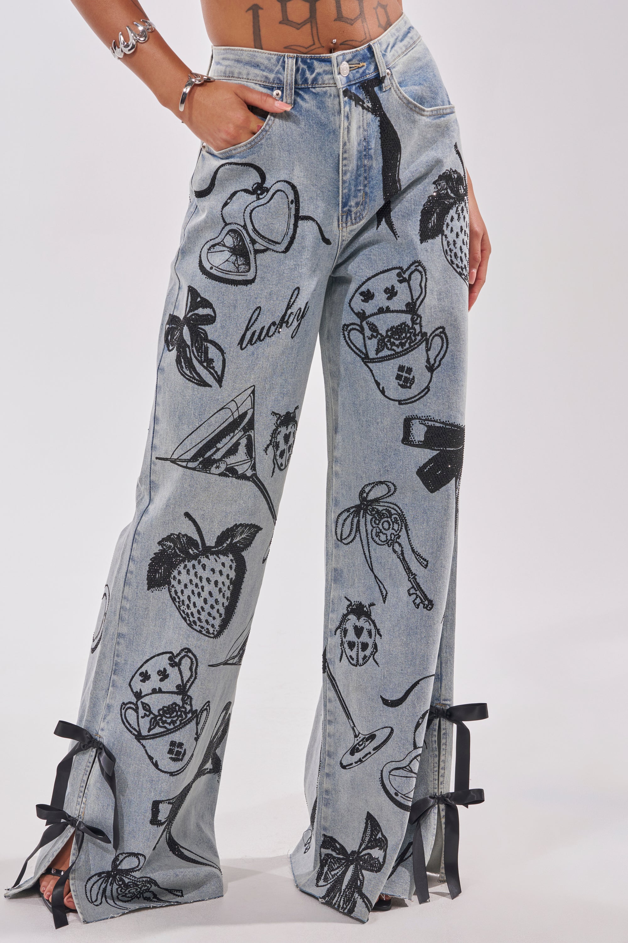 Wearing AFTER HOURS STRAIGHT LEG DENIM—light blue jeans with black graphic prints (strawberries, teapots, keys, hearts, “lucky”), bow accents at the ankles, and a black belt. Tattoos and jewelry add an edgy finish. 2