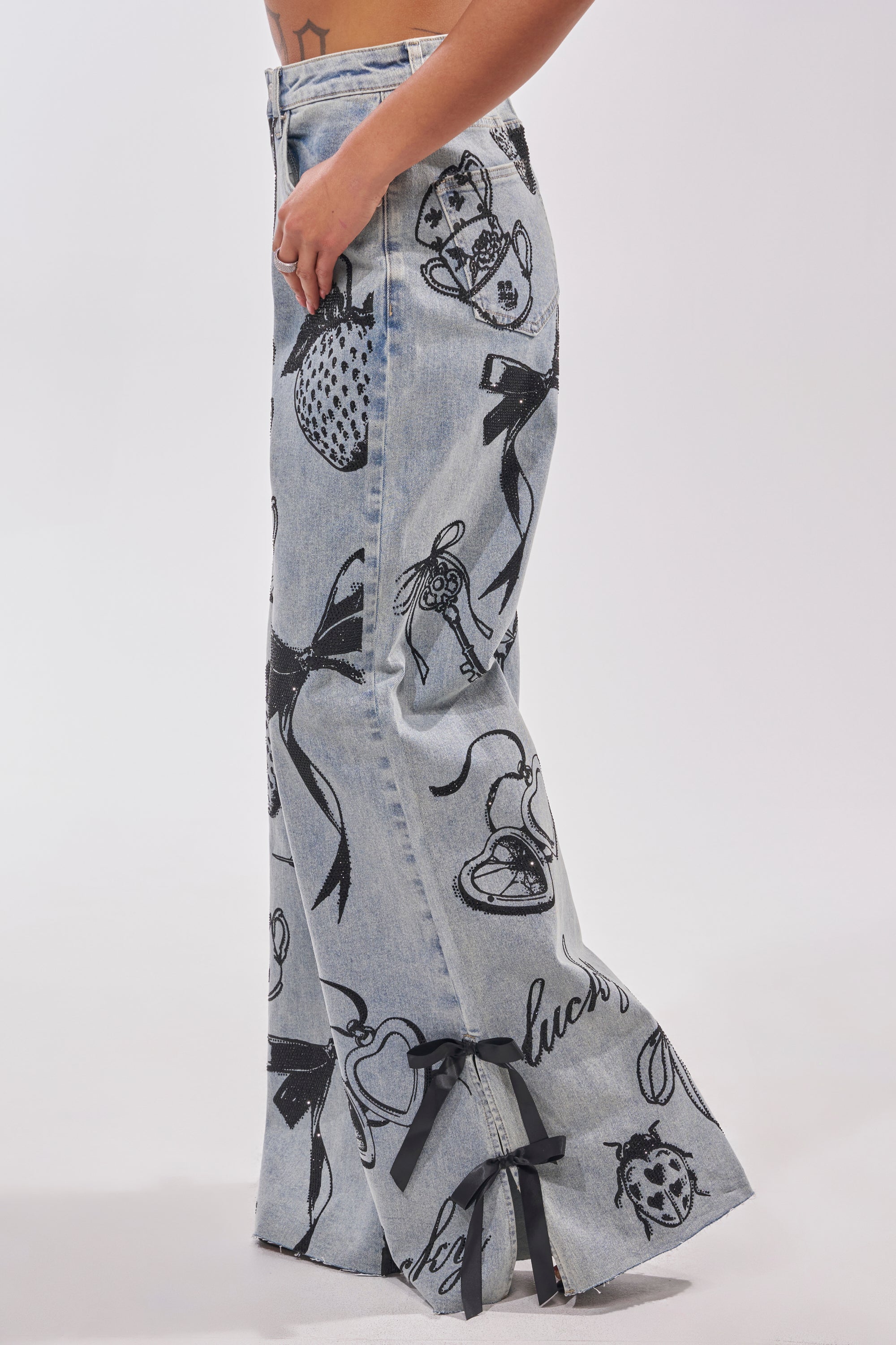 A person models the AFTER HOURS STRAIGHT LEG DENIM, featuring black printed bows, hearts, strawberries, and script. Black ribbon bows accent the hem. Only the lower torso and legs are shown. 3