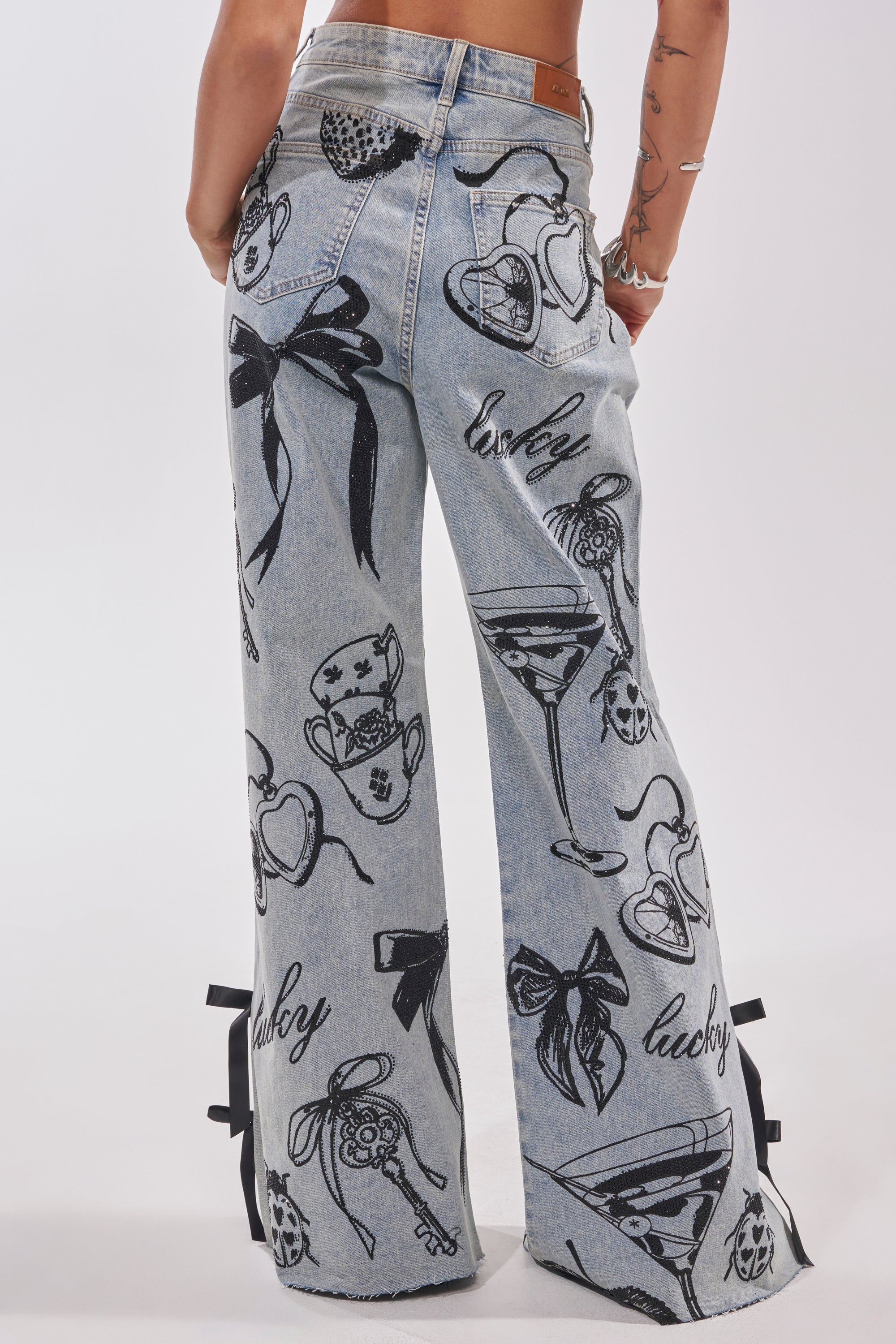 Wearing AFTER HOURS STRAIGHT LEG DENIM in light blue with black heart, bow, pizza slice, martini glass, and "lucky" graphics. Tattoos visible on arms as they stand against a plain background. 4