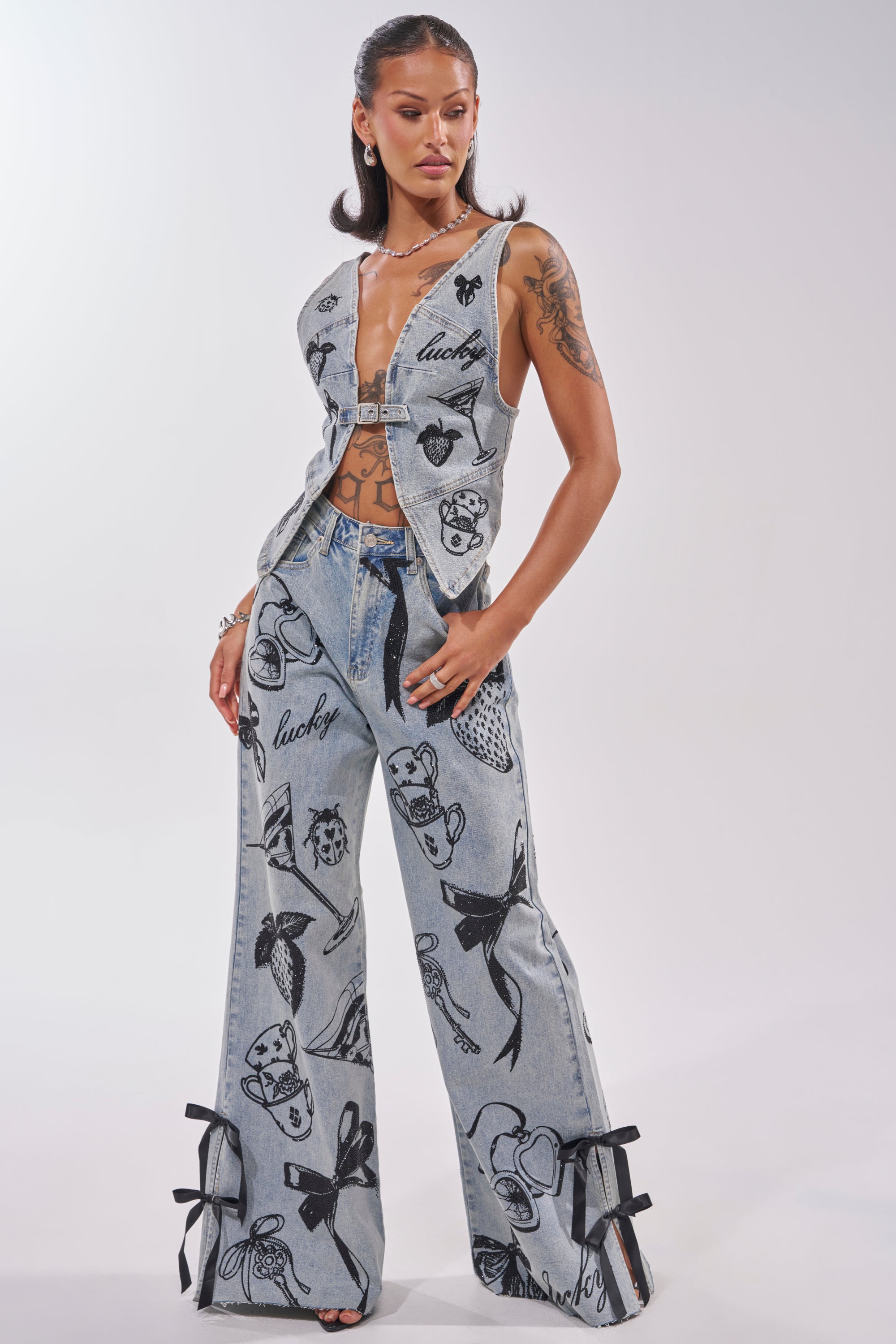 A woman poses in AFTER HOURS STRAIGHT LEG DENIM, featuring bold black prints, "lucky" text, cutout details, and bow accents at the ankles. She pairs it with a sleeveless denim vest and displays tattoos on her arm. 5