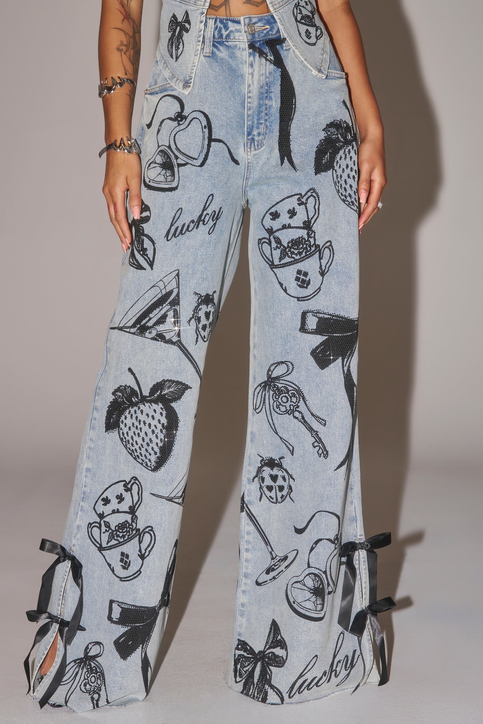 A person is shown from the waist down wearing AFTER HOURS STRAIGHT LEG DENIM—light blue jeans with black ribbon bows at the hems and bold graphic prints of strawberries, teacups, hearts, dice, and the word "lucky. 6